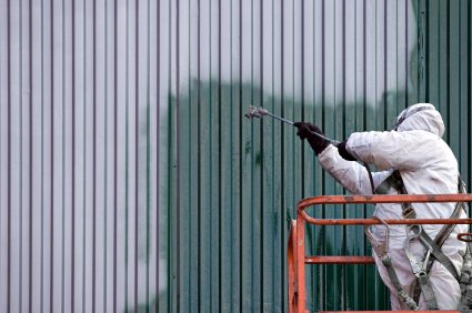 Worker in protective suit spray-painting or cleaning a corrugated metal wall from a lift